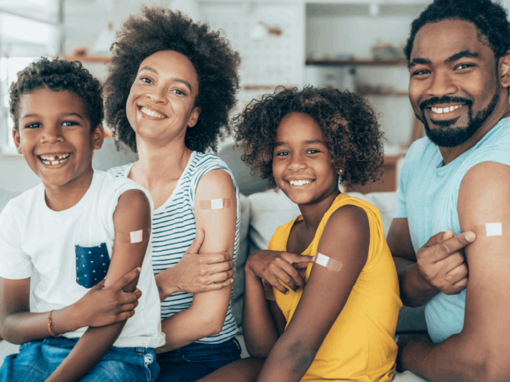 Family of four smiling with a bandage on each arm from their flu shots